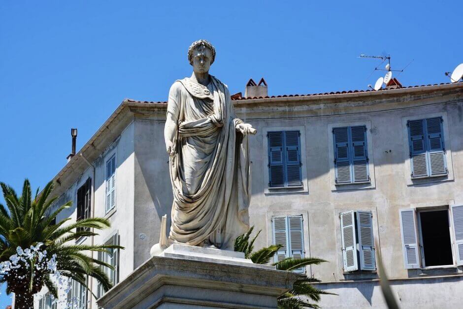 Napoléon représenté en empereur avec sa toge, sur une place ensoleillée de sa ville natale Ajaccio