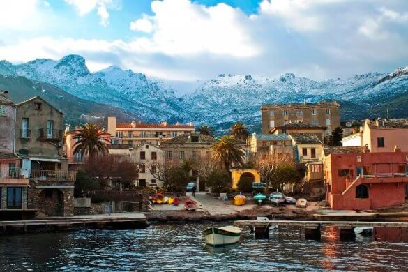 Vue d'un village corse au bord de la mer avec des montagnes enneigées en arrière-plan.