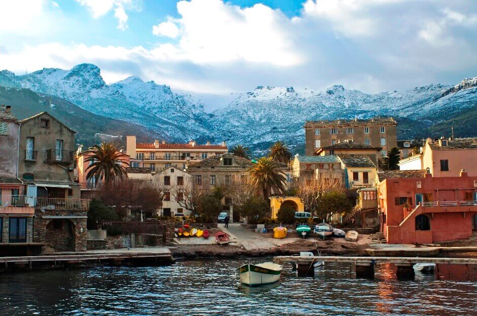 Vue d'un village corse au bord de la mer avec des montagnes enneigées en arrière-plan.