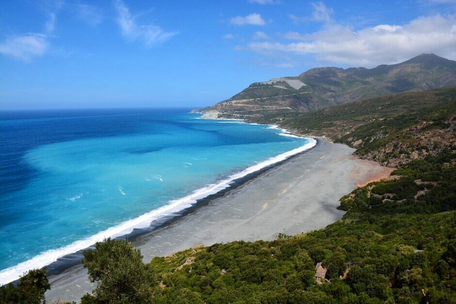 Vue plongeante sur une grande plage grise au pied d'une montagne en Corse.