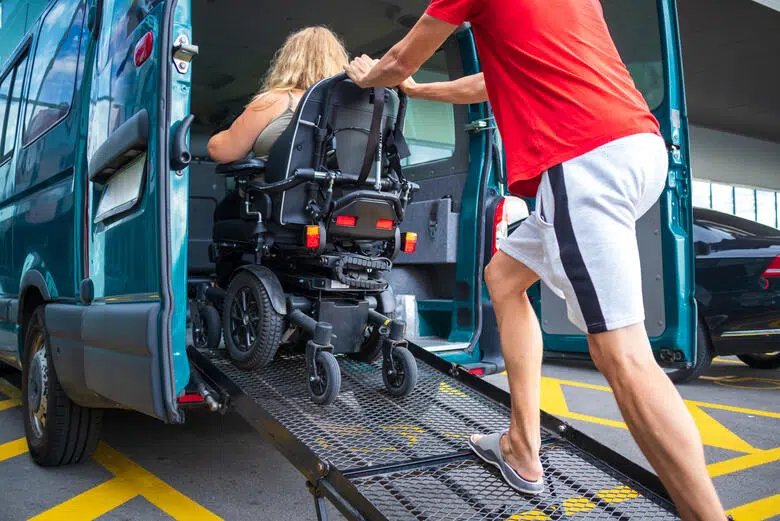 Une femme en fauteuil est assistée pour monter dans un van adapté aux personnes handicapées.